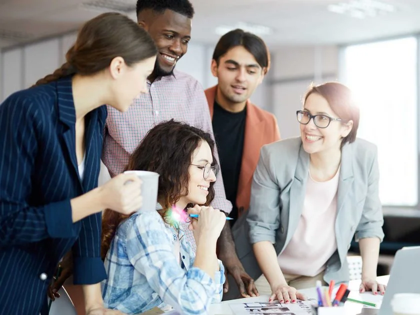 Five young adults, three women and two men, stand around a desk in an office, smiling and talking. One woman holds a mug, while another sits at a laptop. Papers and charts are spread out on the desk.